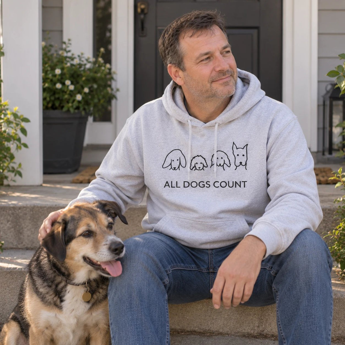 A man on the porch steps with his dog, wearing a grey MoreWoof “All Dogs Count” hoodie