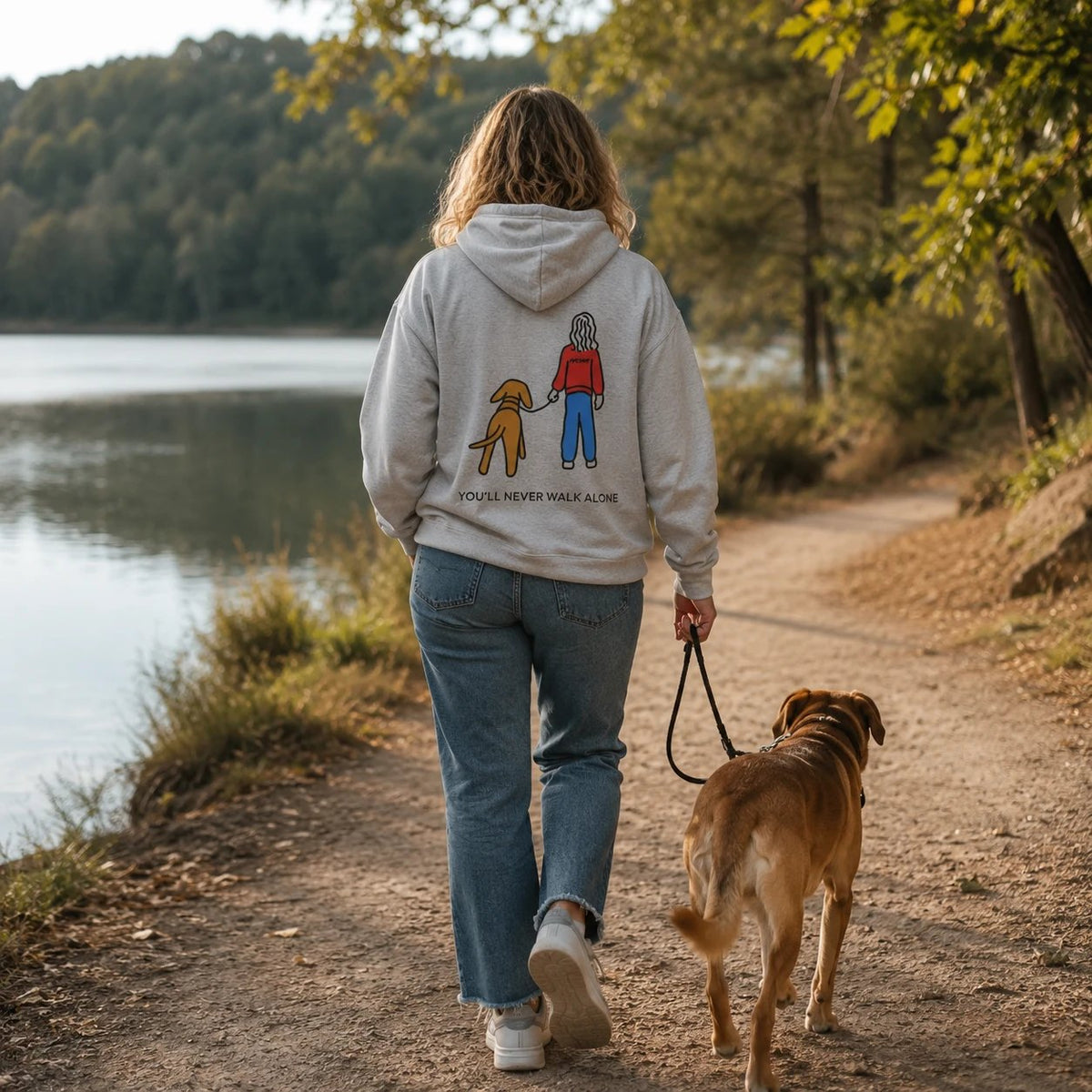 A woman walking her dog by a lake, wearing a MoreWoof “You’ll Never Walk Alone” hoodie